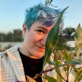 Zarema Khalilova, smiling, with short blue hair, half hidden behind a long green leaf in warm evening light.